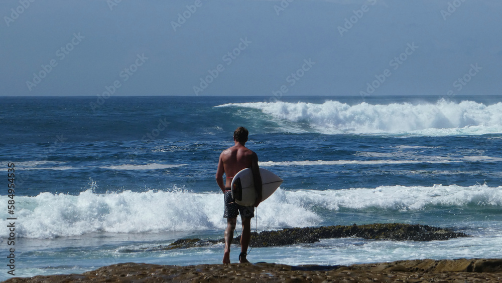 rear view of shirtless man carrying surfboard walking on sandstone rock going for a surf towards huge waves 