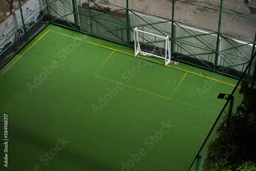 A beautiful, green, empty football playground with a white-colored goalpost, surrounded by green trees, green fence made of net and a street on the background. 
