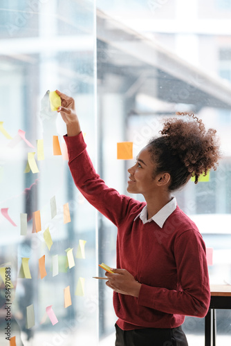African American female employee write down on colorful sticky notes manage list, concentrated biracial woman work on startup brainstorm collaborate plan on stickers on glass wall