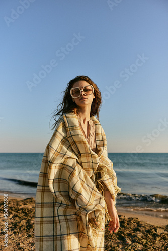 a woman wrapped in a plaid stands on the seashore in bright sunglasses against the background of the sunset sky