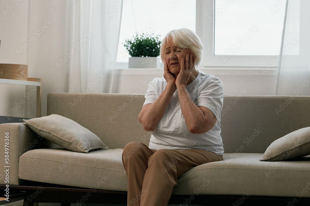 Elderly woman severe pain in the head sitting on the couch, health problems in old age, poor quality of life. Grandmother with gray hair holding her head, migraine and high blood pressure. Elderly woman severe pain in the head sitting on the couch, health problems in old age, poor quality of life. Grandmother with gray hair holding her head, migraine and high blood pressure.