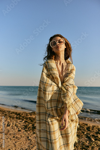 a woman wrapped in a plaid stands on the seashore in bright sunglasses against the background of the sunset sky