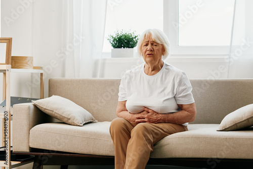 Elderly woman severe abdominal pain sitting on the sofa, health problems in old age, poor quality of life. Grandmother with gray hair holding her stomach, poisoning, problems with stool. © SHOTPRIME STUDIO