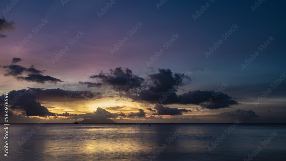Naklejka premium Fantastically beautiful tropical sunset. Purple clouds in the sky, highlighted in pink and gold. Reflection on the calm surface of the ocean. Silhouette of an island and a yacht on the horizon. 