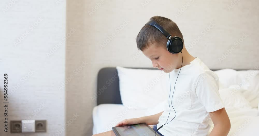 A kid boy in white T-shirt using tablet computer with headphones, siting on bed A kid boy in white T-shirt using tablet computer with headphones, siting on bed