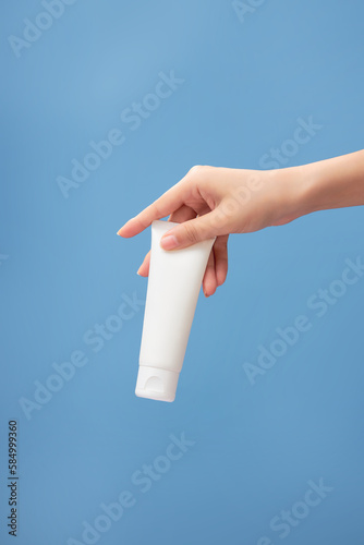 Beautiful female hand holding a tube in white color on blue background. Empty label for product mockup. View from front