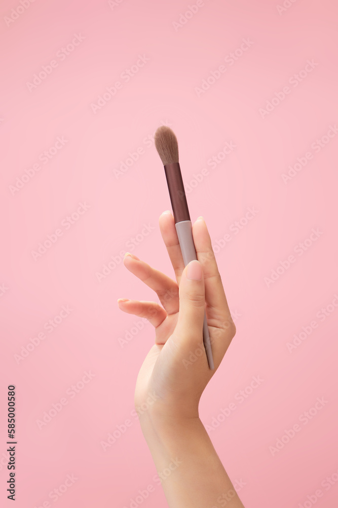 A blending brush is held by a female hand model against light pink background. Makeup product advertising A blending brush is held by a female hand model against light pink background. Makeup product advertising