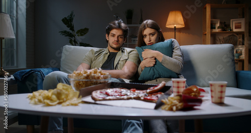 Young caucasian couple spending time in front of tv together  being scared by a horror movie and eating unhealthy food