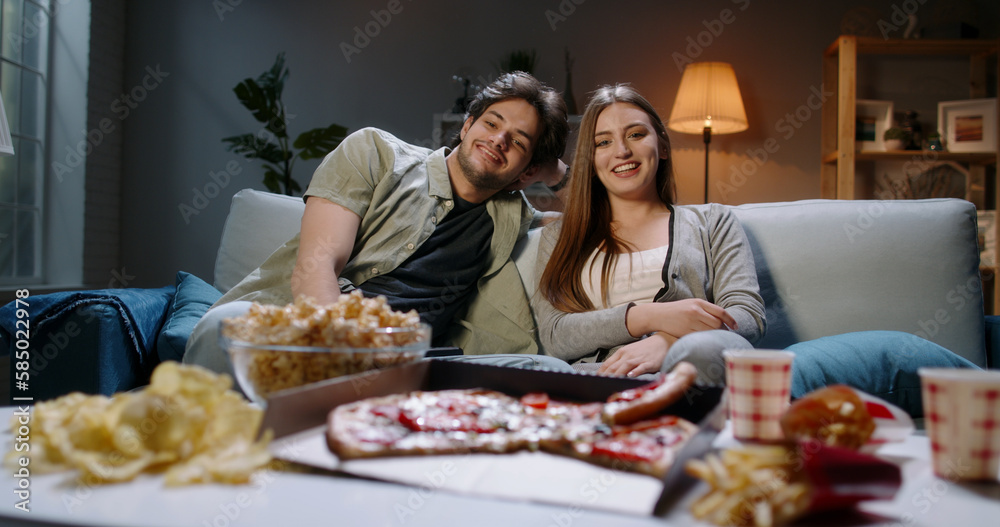 Young happy student caucasian couple chilling together after long day, sitting on couch watch comedies, happily laughing and eating unhealthy food 