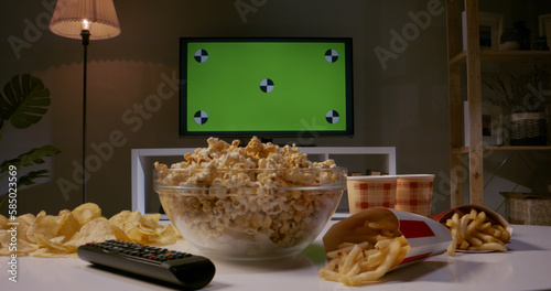 Closeup shot of tv set with green chromakey screen on wall and lots of popcorn, french fries and chips on table ready for watching favourite tv shows 