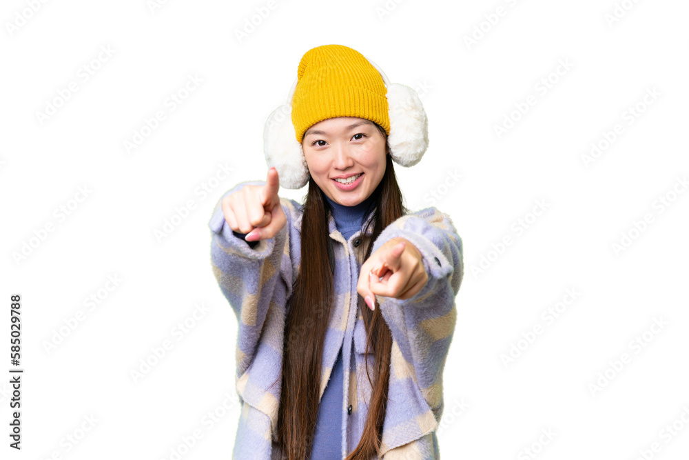 Young Asian woman wearing winter muffs over isolated chroma key background points finger at you while smiling Young Asian woman wearing winter muffs over isolated chroma key background points finger at you while smiling