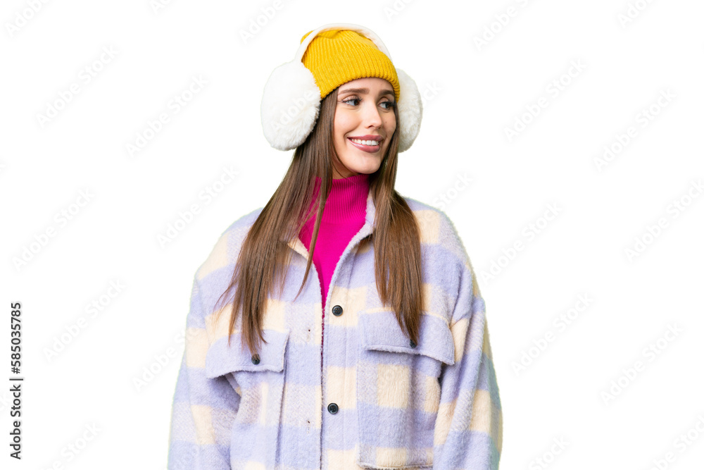 Young woman wearing winter muffs over isolated chroma key background looking side Young woman wearing winter muffs over isolated chroma key background looking side
