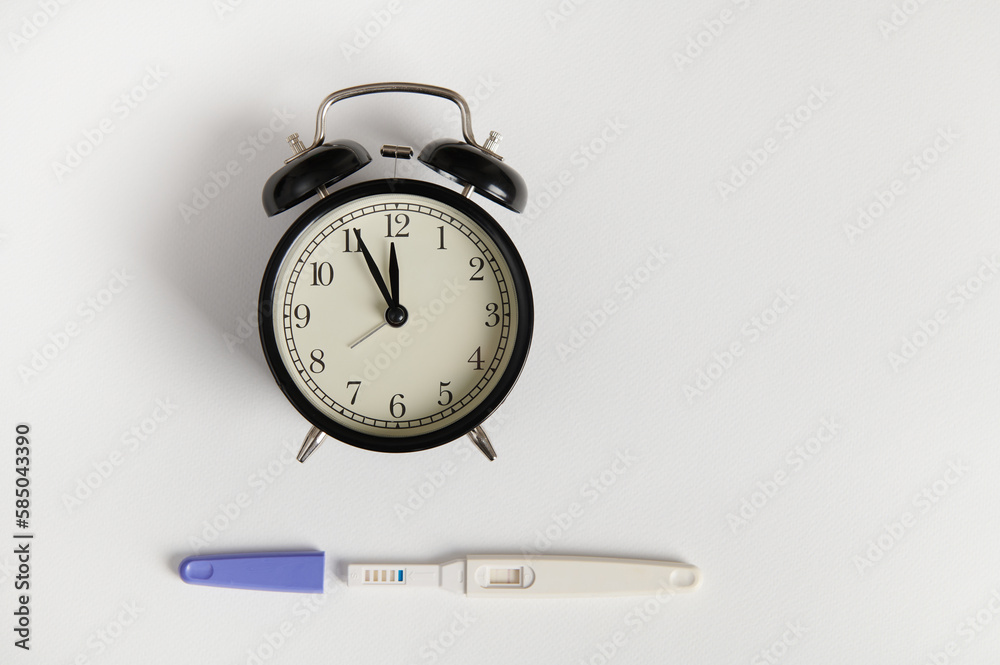 Flat lay with black retro alarm clock and pregnancy test kit showing positive result, isolated white backdrop. Pregnancy, motherhood and gynecological concept. Women's health and fertility. Still life Flat lay with black retro alarm clock and pregnancy test kit showing positive result, isolated white backdrop. Pregnancy, motherhood and gynecological concept. Women's health and fertility. Still life