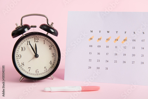 Still life with a black vintage alarm clock, calendar with days of the last menstruation marked and positive inkjet pregnancy test on pink pastel background. Rapid diagnosis of pregnancy at home photo