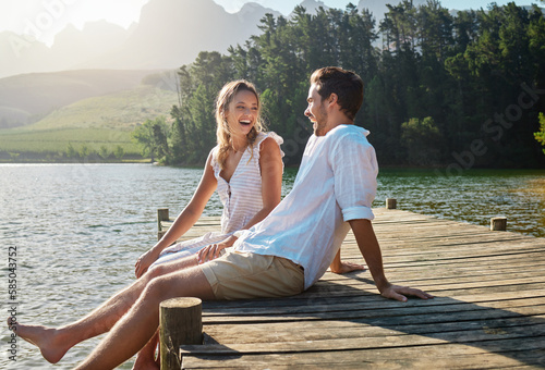 Love, laugh and pier with couple at lake for bonding, romance and affectionate date. Nature, travel and holiday with man and woman sitting on boardwalk in countryside for happy, summer and vacation photo