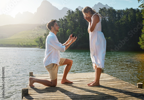 Man, woman and marriage proposal by lake on vacation with surprise, wow or happiness in sunshine. Couple, engagement and offer ring in nature for romance, love and happy on holiday in summer by water photo