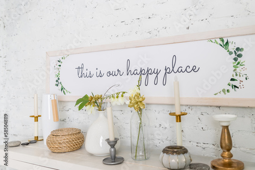 Frame with text HAPPY place, white ceramic vase withplants,flowers, candles on a wooden table or shelf on a background of light brick wall. Stylish spring home interior decor.Vintage style photo