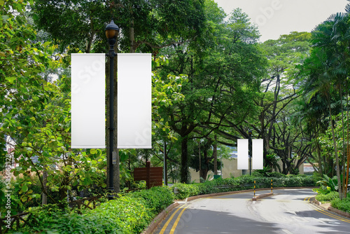 Blank vertical advertising banners on street lampposts; double hanging posters by the road, against lush green trees and plants. For OOH out of home template mock up