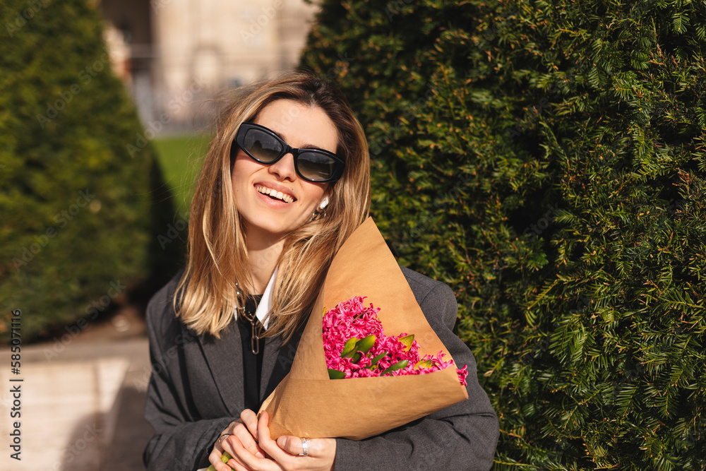 Young beautiful smiling hipster woman in trendy spring outfit. Sexy carefree woman posing on the park background in jacket, sunglasses. Positive model sitting outdoors. Holding bouquet of pink flowers Young beautiful smiling hipster woman in trendy spring outfit. Sexy carefree woman posing on the park background in jacket, sunglasses. Positive model sitting outdoors. Holding bouquet of pink flowers