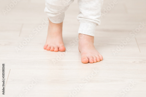 Little child legs in white trousers on light wooden floor background. Barefoot closeup. Front view.