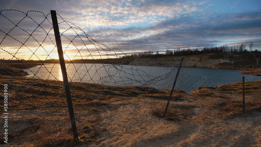 Barbed wire and fence against backdrop of majestic sunset and lake: quarry, sand, picturesque sky. Barbed wire and fence against backdrop of majestic sunset and lake: quarry, sand, picturesque sky.