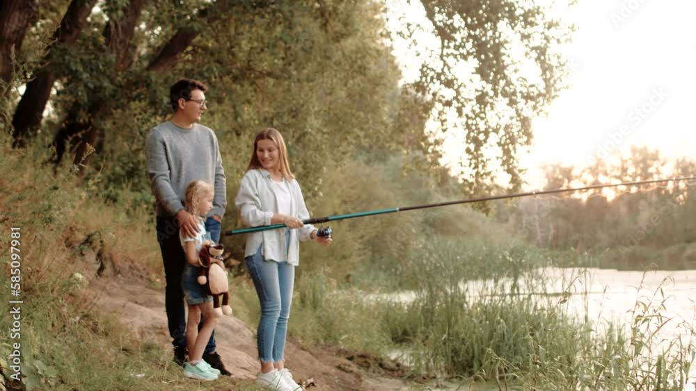 Father, mother and daughter are fishing on riverbank in summer throwing fishing rod into water, side view. Happy family is relaxing at nature, angling on river Father, mother and daughter are fishing on riverbank in summer throwing fishing rod into water, side view. Happy family is relaxing at nature, angling on river