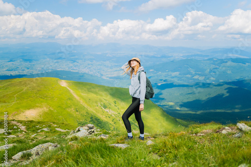 Portrait of a happy woman hiker standing on the slope of mountain ridge against mountains, blue cloudy sky on background. The woman is happy and looking at camera. photo
