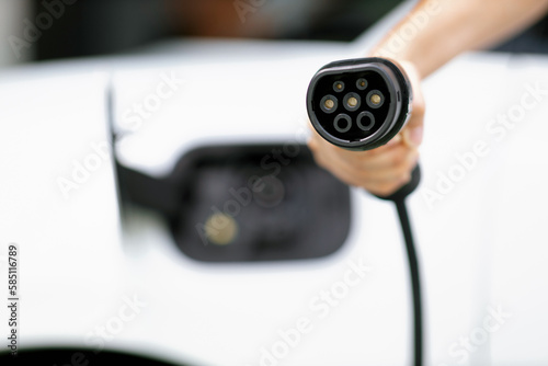 Progressive new alternative clean energy vehicle and electric charging device concept. Focus closeup hand holding and pointing EV charger plug at camear from electric charging station at camera photo