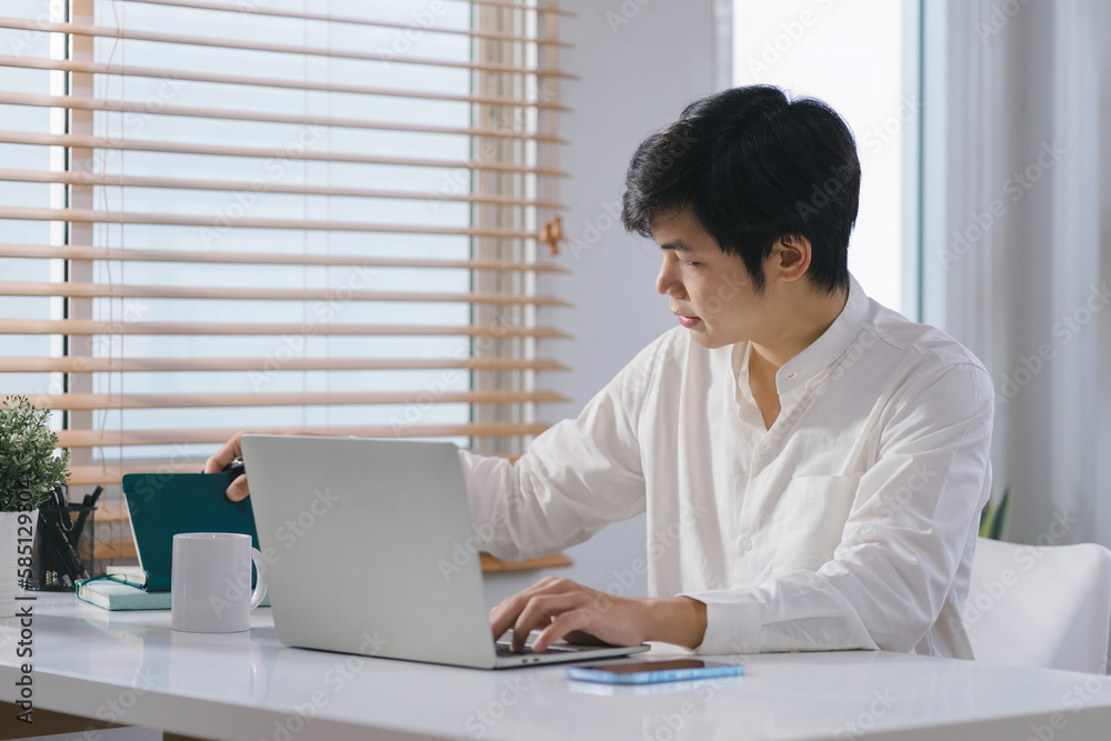 Fototapeta premium Happy and relaxed millennial Asian businessman is working remotely from his home office with his laptop.