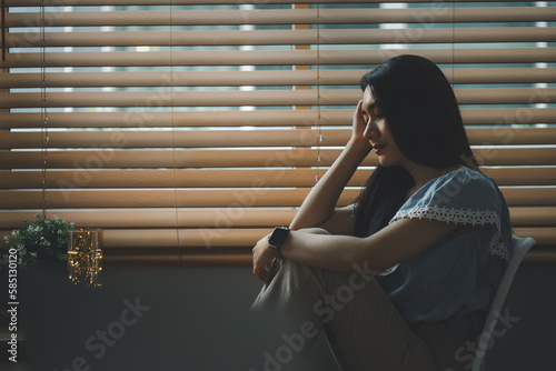 Sad and depressed young woman sitting nere window in the living room, looking outside with a sad expression, conveying feelings of exhaustion, loneliness, and unhappiness. photo