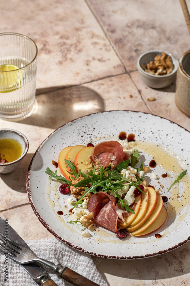 Salad of arugula and prosciutto or ham , sliced apple, cheese, walnuts on rustic plate in composition with cutlery and water glass on pink tile background. Copy space