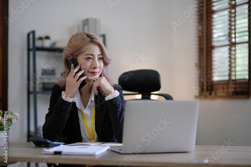 Young Asian businesswoman sit with her laptop computer happily talking on the phone with her customer explaining the detail in the office.