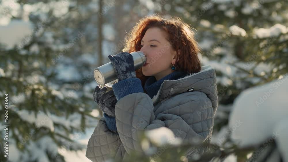 Close-up shot of a beautiful red-haired woman standing in the middle of snow-covered trees and drinking tea from a thermos. A woman walking in a snowy park in the morning drinks hot tea from a thermos Close-up shot of a beautiful red-haired woman standing in the middle of snow-covered trees and drinking tea from a thermos. A woman walking in a snowy park in the morning drinks hot tea from a thermos