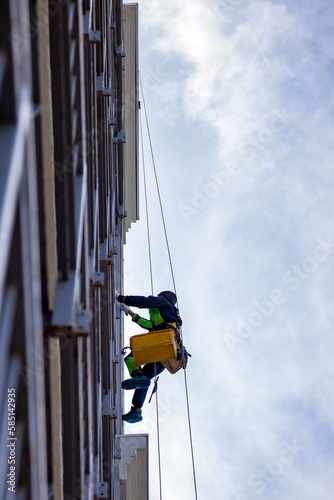 Occupational industrial mountaineering worker washing exterior facade glazing building at blue sky background, hangs over house. Rope access laborer climbing on wall of skyscraper. Copy ad text space