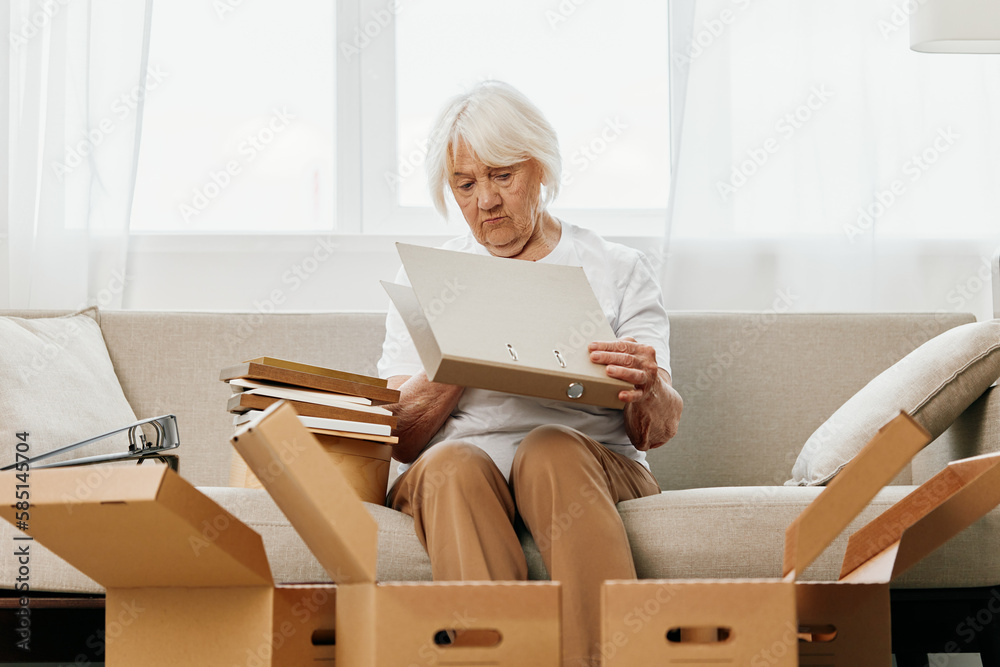 elderly woman sits on a sofa at home with boxes. collecting things with memories albums with photos and photo frames moving to a new place cleaning things and a happy smile. Lifestyle retirement. elderly woman sits on a sofa at home with boxes. collecting things with memories albums with photos and photo frames moving to a new place cleaning things and a happy smile. Lifestyle retirement.