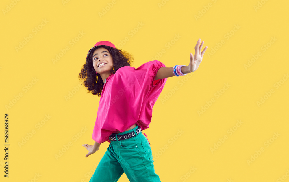 Happy child dancing. Cheerful little African American girl wearing loose oversized fuschia top and green jeans with trendy eyelet belt dancing isolated on solid yellow background. Kids fashion concept - obrazy, fototapety, plakaty 