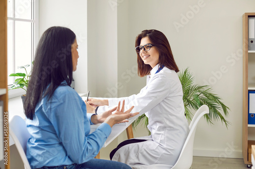 Friendly smiling doctor is listening to his female patient who is talking about her health complaints. Female doctor conducts consultations for patients at her workplace in office in medical clinic.