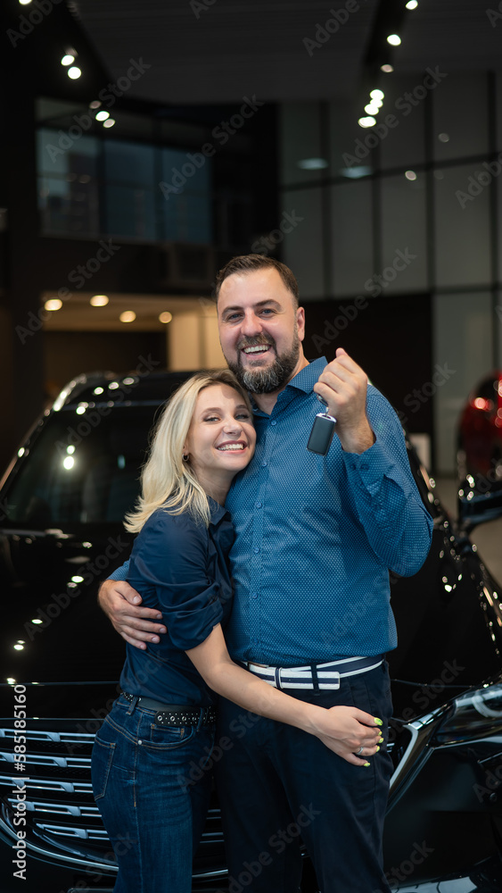 Happy caucasian couple hugging and man holding new car keys in car showroom.