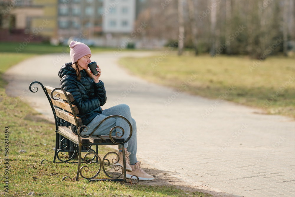 Street portrait of alone woman with cup of coffee seated on wooden bench at urban park in springtime Street portrait of alone woman with cup of coffee seated on wooden bench at urban park in springtime