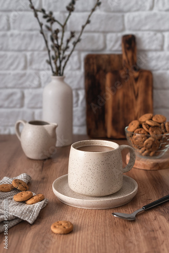 A ceramic cup with coffee, a milk jug, a glass bowl with cookies and a vase with branches on a wooden kitchen table. Still life in the style of Scandi. Home comfort.