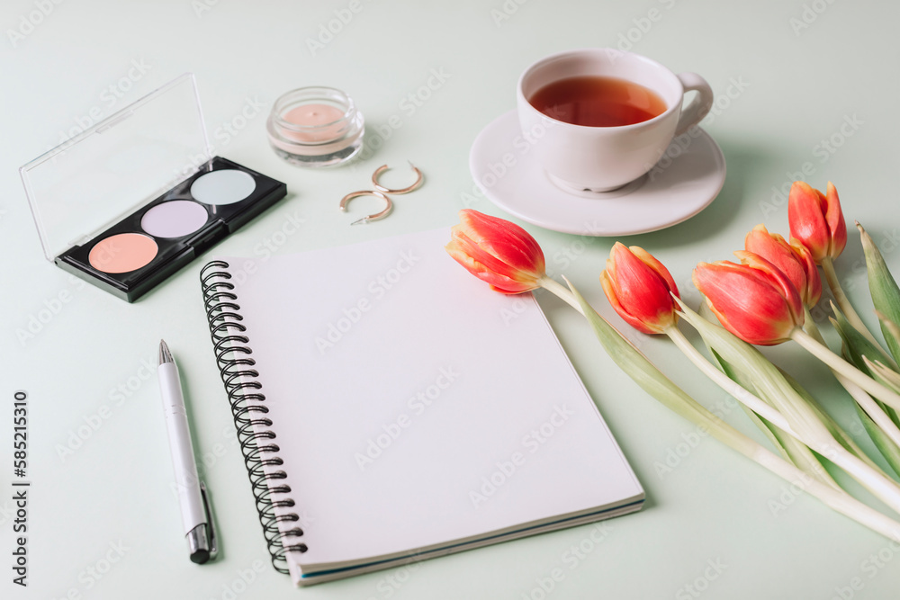 Blank notepad, cup of tea, makeup cosmetics and bouquet of red tulips on light green background. Top view, mockup. Women's day or Mother's day concept Blank notepad, cup of tea, makeup cosmetics and bouquet of red tulips on light green background. Top view, mockup. Women's day or Mother's day concept