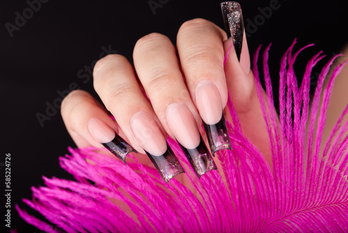 Hand with long artificial french manicured nails colored with black nail polish with glitter and gemstones decoration and pink feather. Black background.