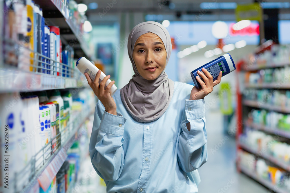 A young Muslim woman in a hijab is standing in a supermarket in the household chemicals department and holding two shampoos. He looks confusedly at the camera, chooses, advertises, shows.
