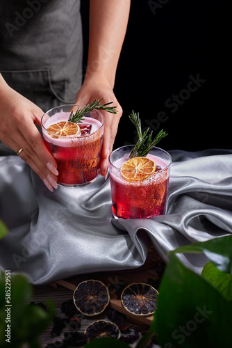 Red drink cocktail of rum and mezcal with lime and cherry juice garnished with dried fruit and green rosemary placed by two female hands