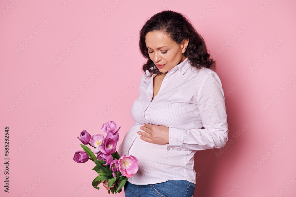 Attractive middle-aged pregnant woman touching her tummy, feels first baby kicks, holding a beautiful bouquet of tulips on isolated pink background. Happy mothers Day. Pregnancy and maternity concept Attractive middle-aged pregnant woman touching her tummy, feels first baby kicks, holding a beautiful bouquet of tulips on isolated pink background. Happy mothers Day. Pregnancy and maternity concept