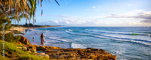 A beautiful girl in a dress stands on a rocky cliff looking at the famous Coolum beach and the Pacific Ocean and surfers catching big waves. Panorama from Sunshine Coast, Queensland, Australia
