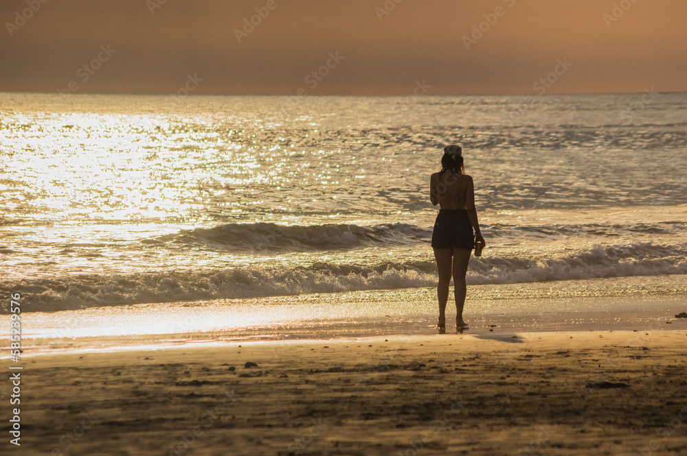 Silhouette of a girl watching the sunset on the beach near the sea and a drink in her hand with a background of the sea reflecting sunlight