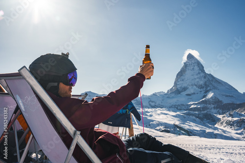 Quadro su tela Young snowboarder cheering with a beer after skiing day in a bar or a cafe at the Zermatt ski resort in Swiss Alps with a fabulous view of the Matterhorn