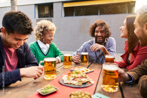 Multiracial friends having fun drinking beer together at terrace bar. Young people laughing at restaurant cafeteria. Happy guys and girls. High quality photo