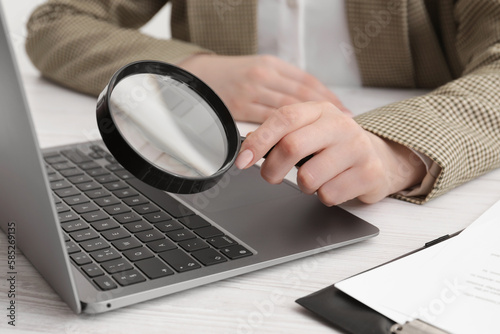 Woman holding magnifier near laptop at white wooden table, closeup. Online searching concept
